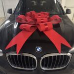 Голяма червена декоративна панделка, поставена върху автомобил BMW, подходяща за подарък, изненада или специален повод.