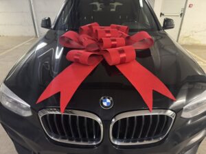 Голяма червена декоративна панделка, поставена върху автомобил BMW, подходяща за подарък, изненада или специален повод.